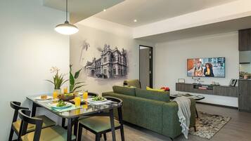 A modern living room with a dining area, a green sofa, a flat-screen TV, and a kitchen area with a built-in oven.