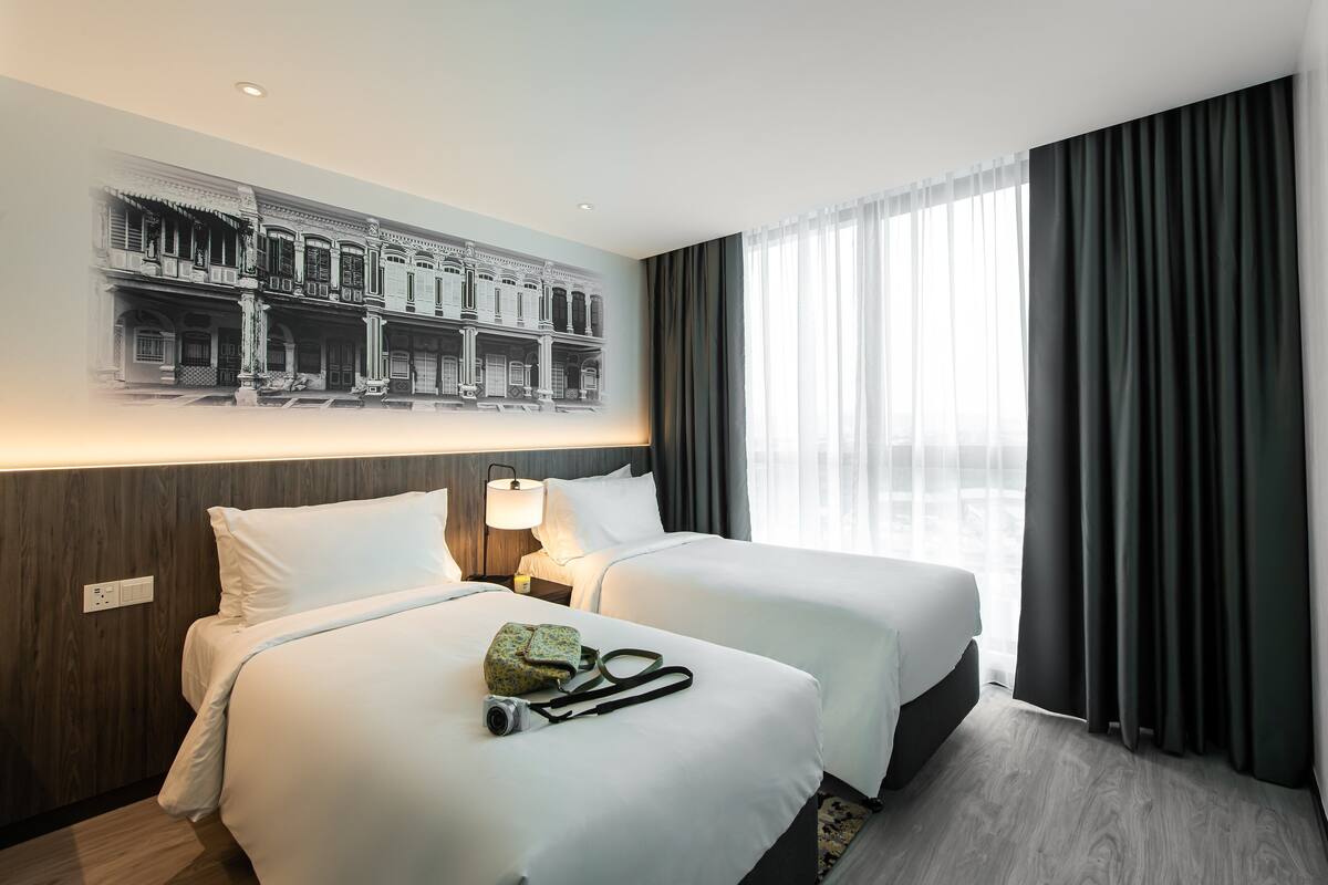 A hotel room with two beds, a large window with curtains, a wall-mounted light, and a wall art depicting a building.