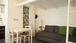 A compact living space with a white table and chairs, a grey sofa with green cushions, and a small round table with a potted plant. - Dependance in Villa con Piscina by Wonderful Italy (Siracusa)