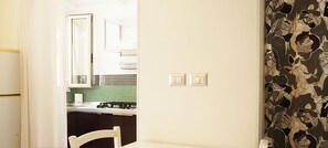 A small, bright kitchen with a white countertop, a stove, and a sink. There is a white chair and a table in the foreground. The walls are plain with a floral wallpaper on one side.