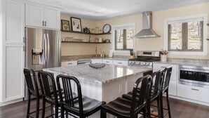 A modern kitchen with white cabinets, a large island with bar stools, stainless steel appliances, and a window overlooking a garden. - Modern Retreat with Apartment and Mountain Views! (Waitsfield)