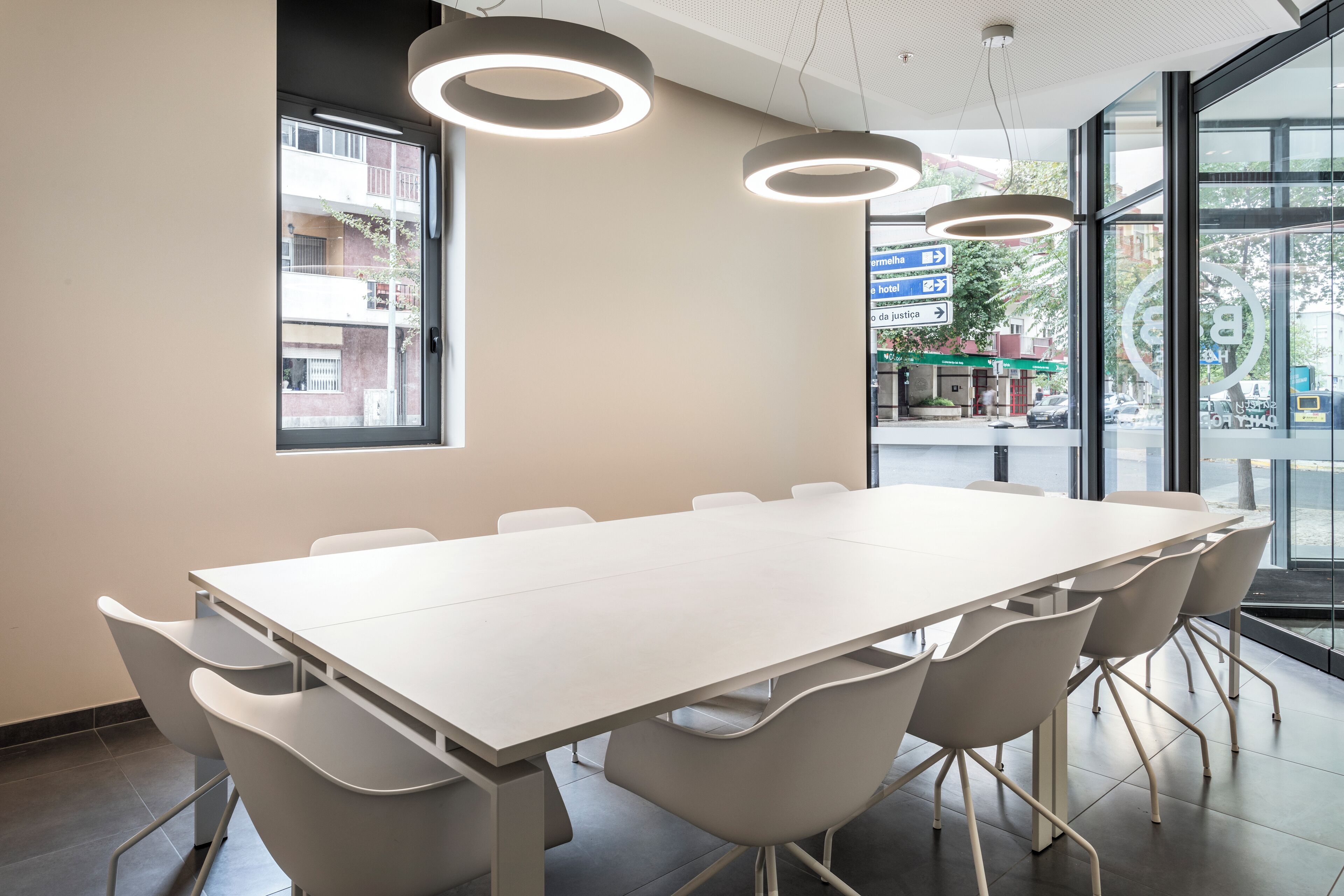 Uma sala de conferências com uma mesa comprida e cadeiras modernas, amplas janelas com vista para a rua e luminárias circulares no teto.