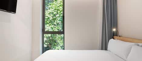 A modern hotel room with a large window, a flat-screen TV mounted on the wall, and a neatly made bed with white linens.