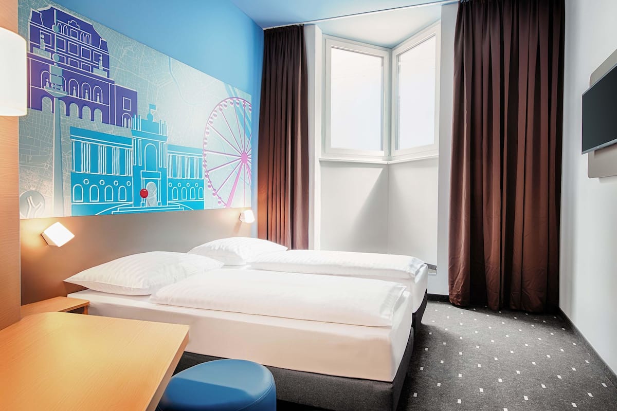 A hotel room with a bed, a desk, a chair, a window with curtains, and a wall mural.