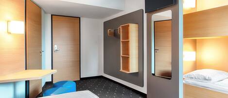 Una habitación de hotel con literas, un escritorio con silla, un armario, un espejo y estante en la pared.