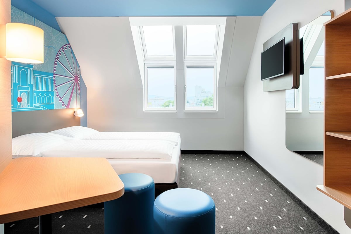 A modern hotel room with a bed, a desk, two blue ottomans, a wall-mounted TV, and large windows with city views.