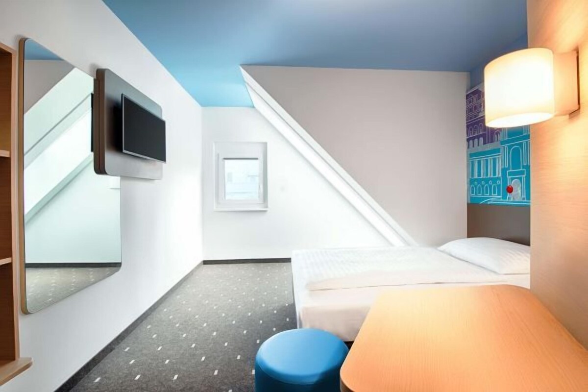 A hotel room with a bed, a desk, a chair, a television, a window, and a staircase.