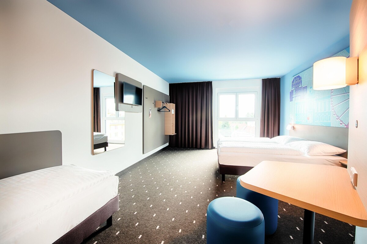 Ein Hotelzimmer mit zwei Betten, einem Schreibtisch, einem Stuhl, einem Fernseher und einem Fenster mit Vorhängen.