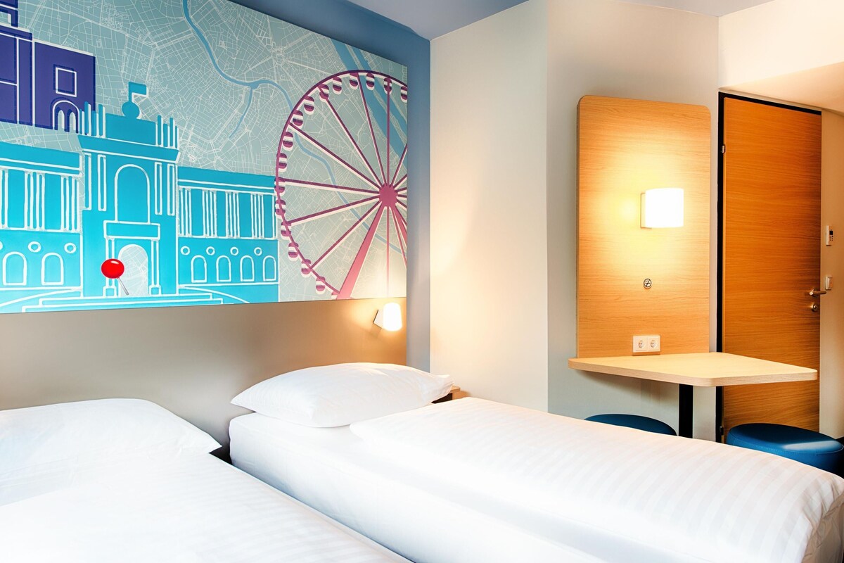 Ein Hotelzimmer mit zwei Betten, einer Wandkarte, einer Holztür und einem kleinen Tisch mit einer Lampe.