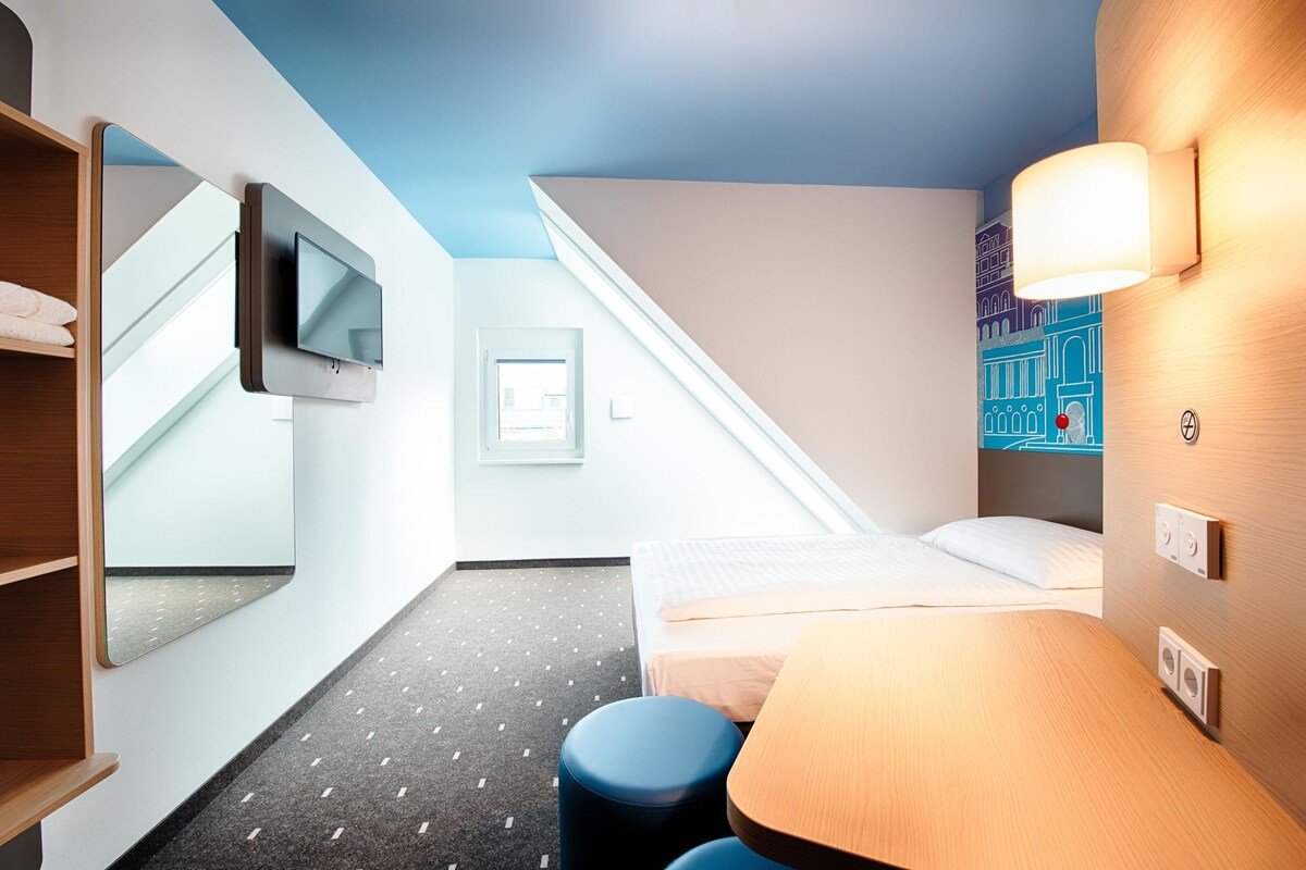 Ein Hotelzimmer mit Bett, Schreibtisch, an der Wand montiertem Fernseher, Badezimmer mit Dusche und einer Treppe, die in einen oberen Bereich führt.
