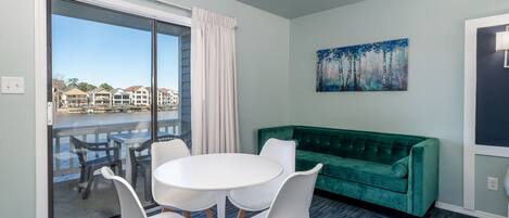 Quarto com uma grande porta de vidro deslizante que dá para uma varanda, um sofá de veludo verde, uma mesa branca redonda com quatro cadeiras e um quadro na parede.