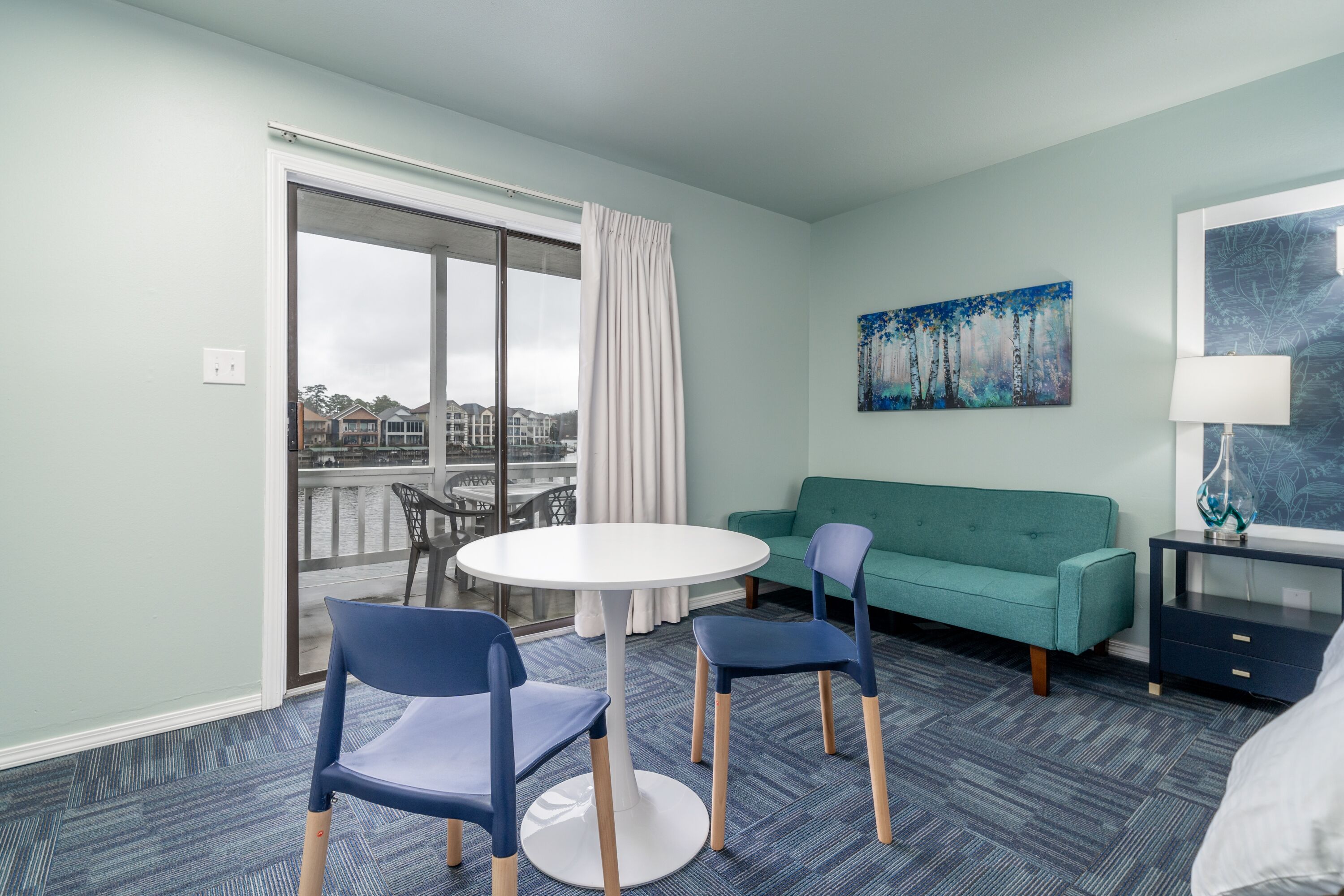 Hotellihuone, jossa on parveke, pyöreä pöytä tuoleineen, vihreä sohva, yöpöytä ja taulu seinällä.