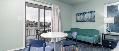 Hotellihuone, jossa on parveke, pyöreä pöytä tuoleineen, vihreä sohva, yöpöytä ja taulu seinällä.