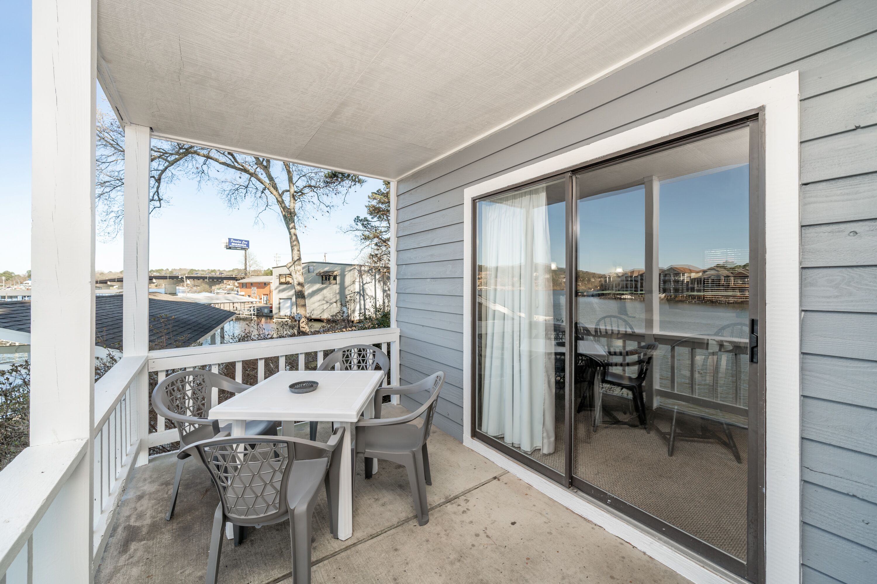 A covered outdoor patio with a table and chairs, a sliding glass door, and a view of a bridge and buildings in the background.