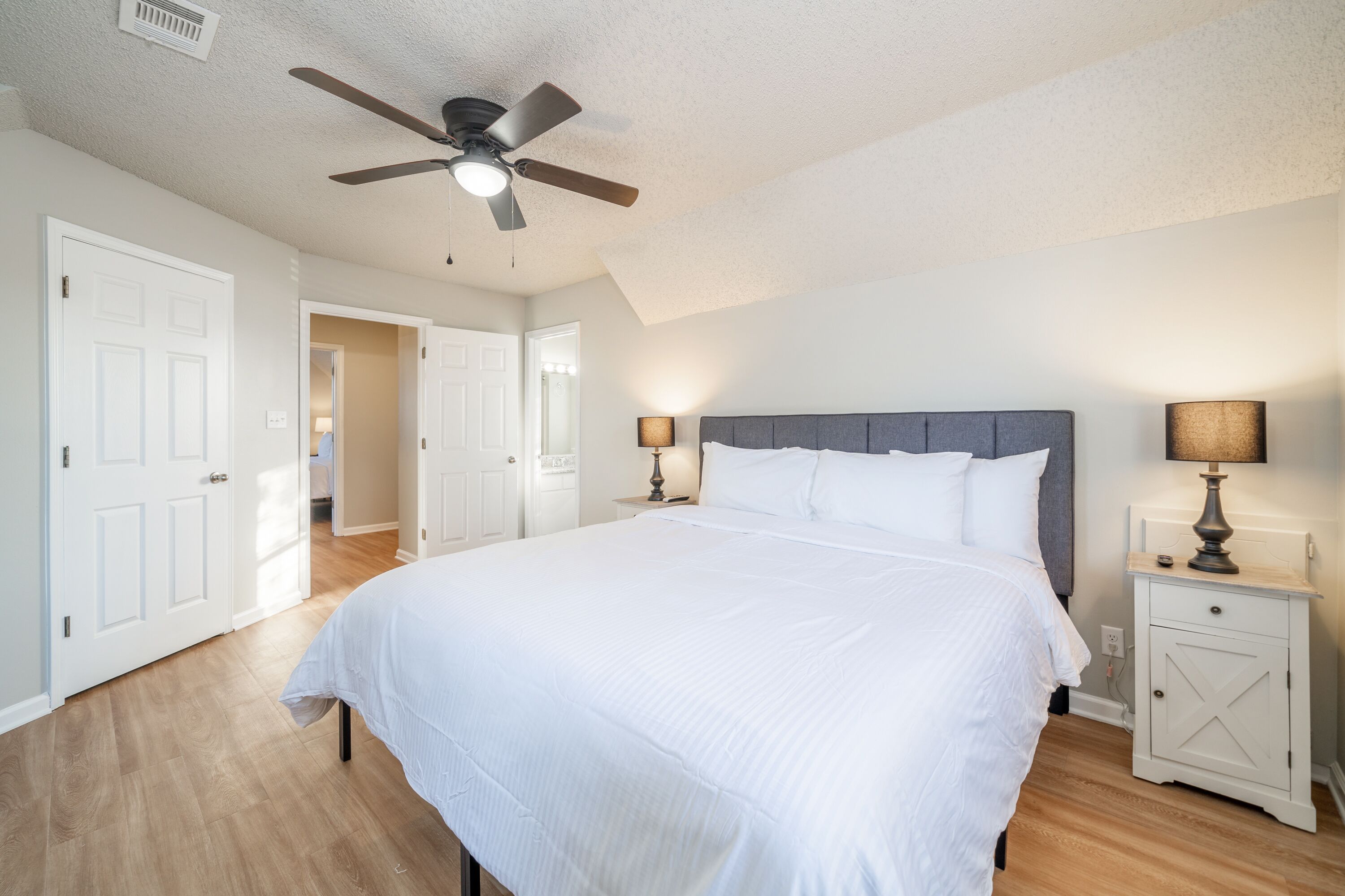 A bedroom with a large bed, two bedside tables with lamps, a ceiling fan, and a door leading to another room.