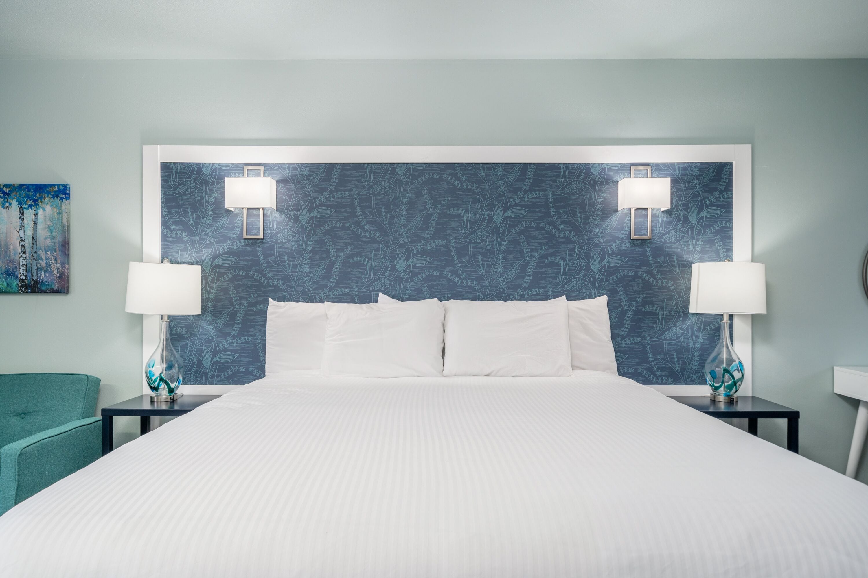 Una camera d'hotel con un letto grande, due comodini con lampade, una poltrona verde acqua e una parete decorata con un motivo a foglia.