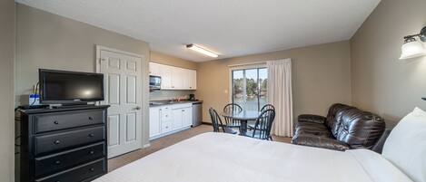 Una habitación de hotel con una cama grande, televisión, una mesa de comedor con sillas, una zona de cocina y un balcón con cortinas.