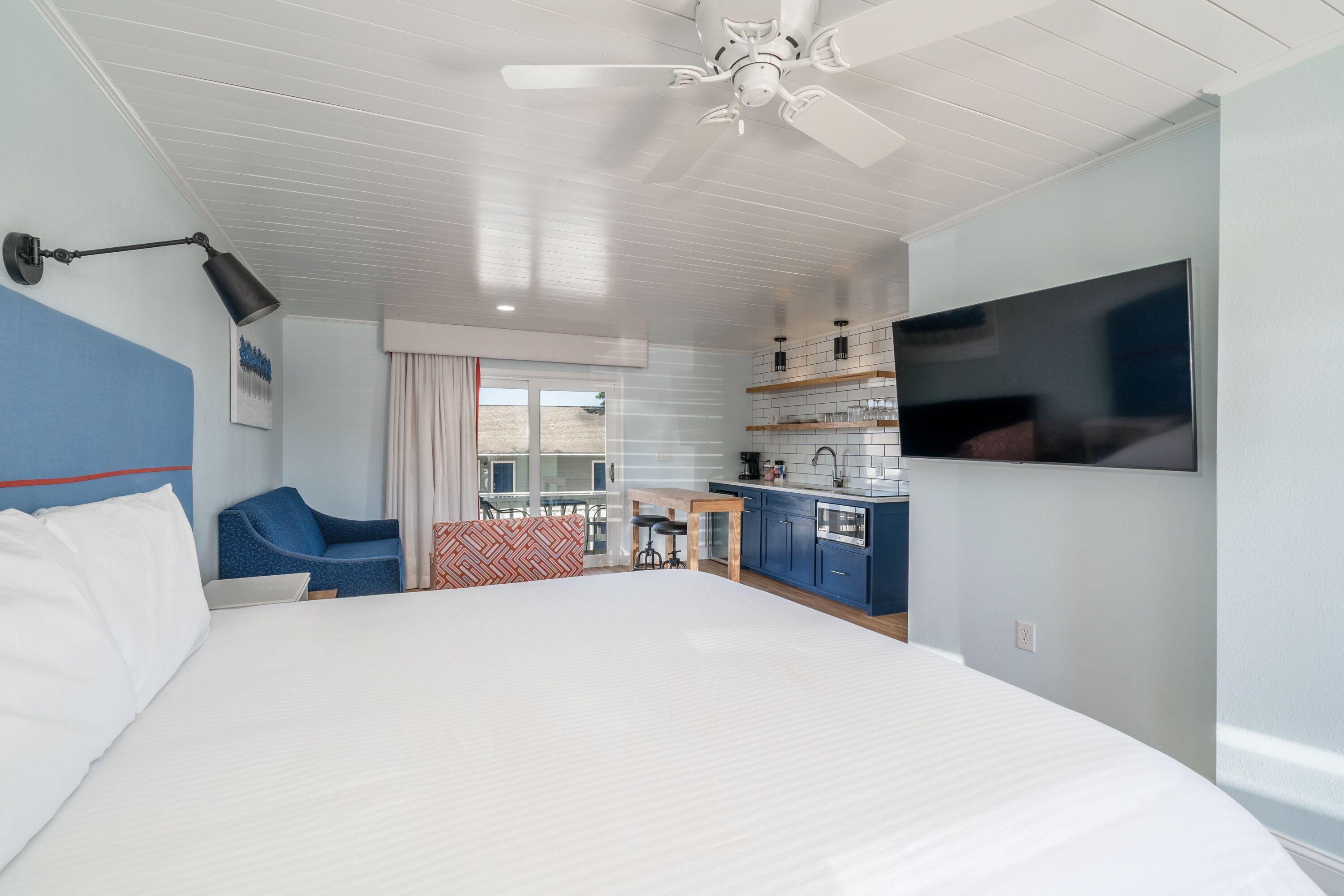 A modern hotel room with a large bed, a desk, a chair, a TV, and a kitchenette.