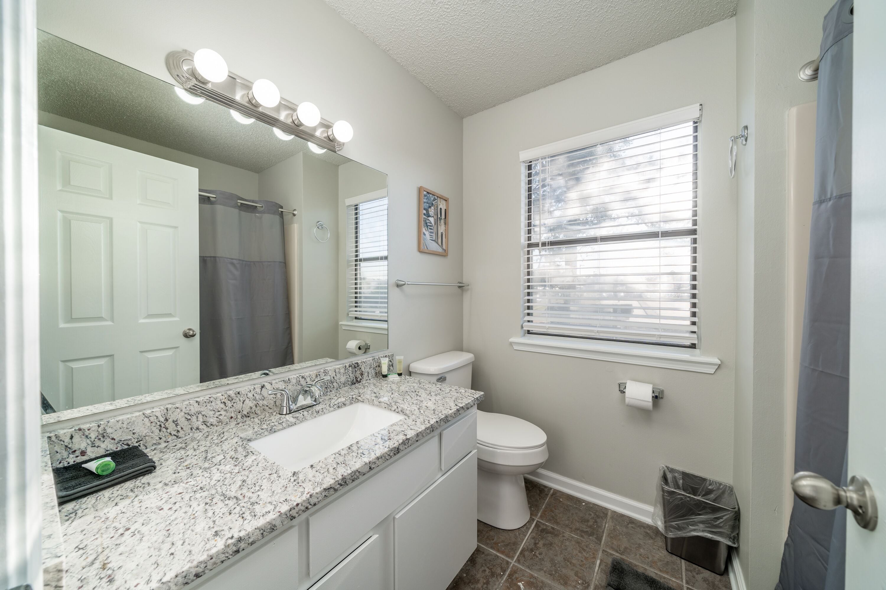 A bathroom with a white sink, vanity, and toilet. It has a large mirror with lights, a window with blinds, and a shower curtain.