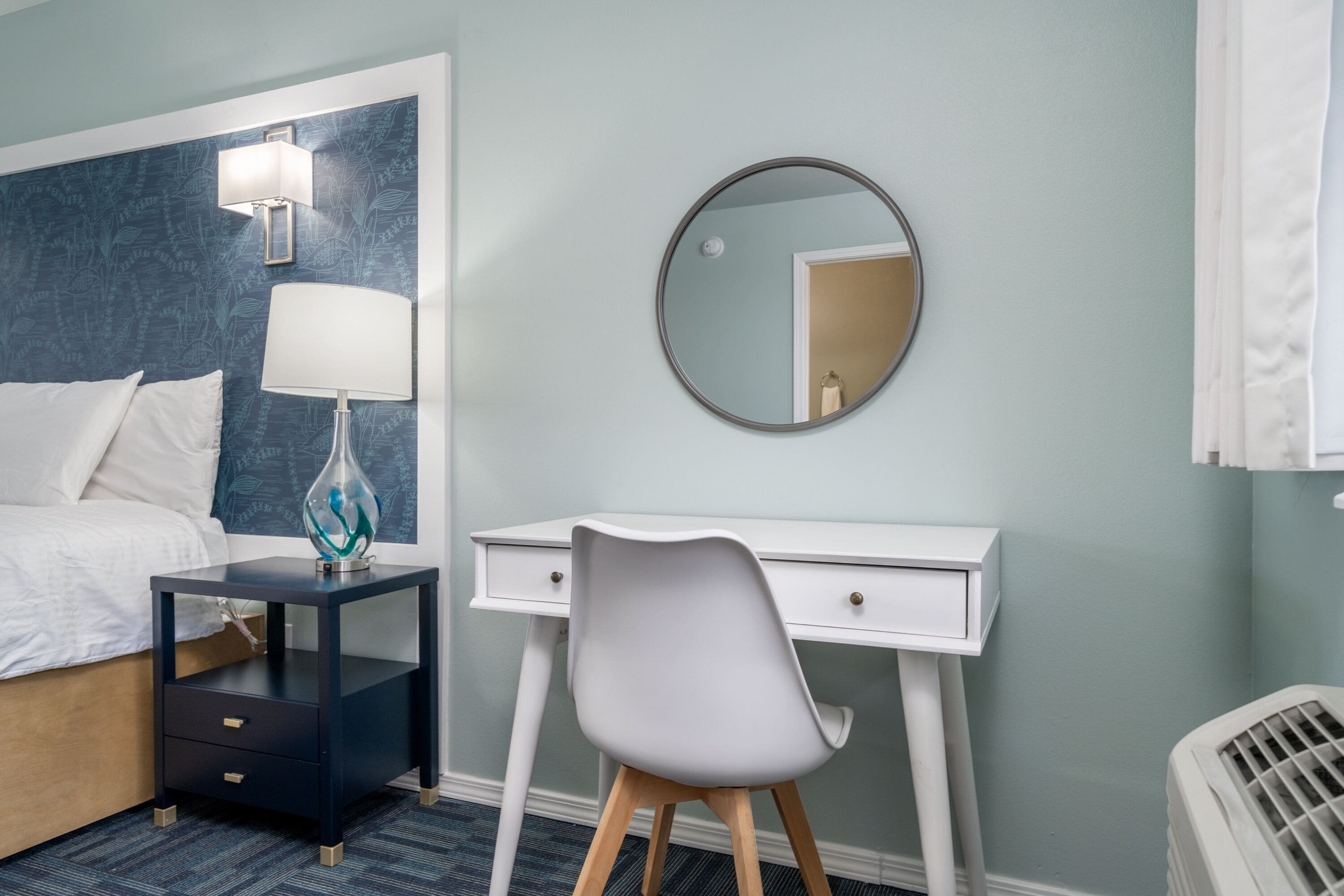 Habitación de hotel con cama, escritorio, silla, mesita de noche, lámpara, espejo y radiador.