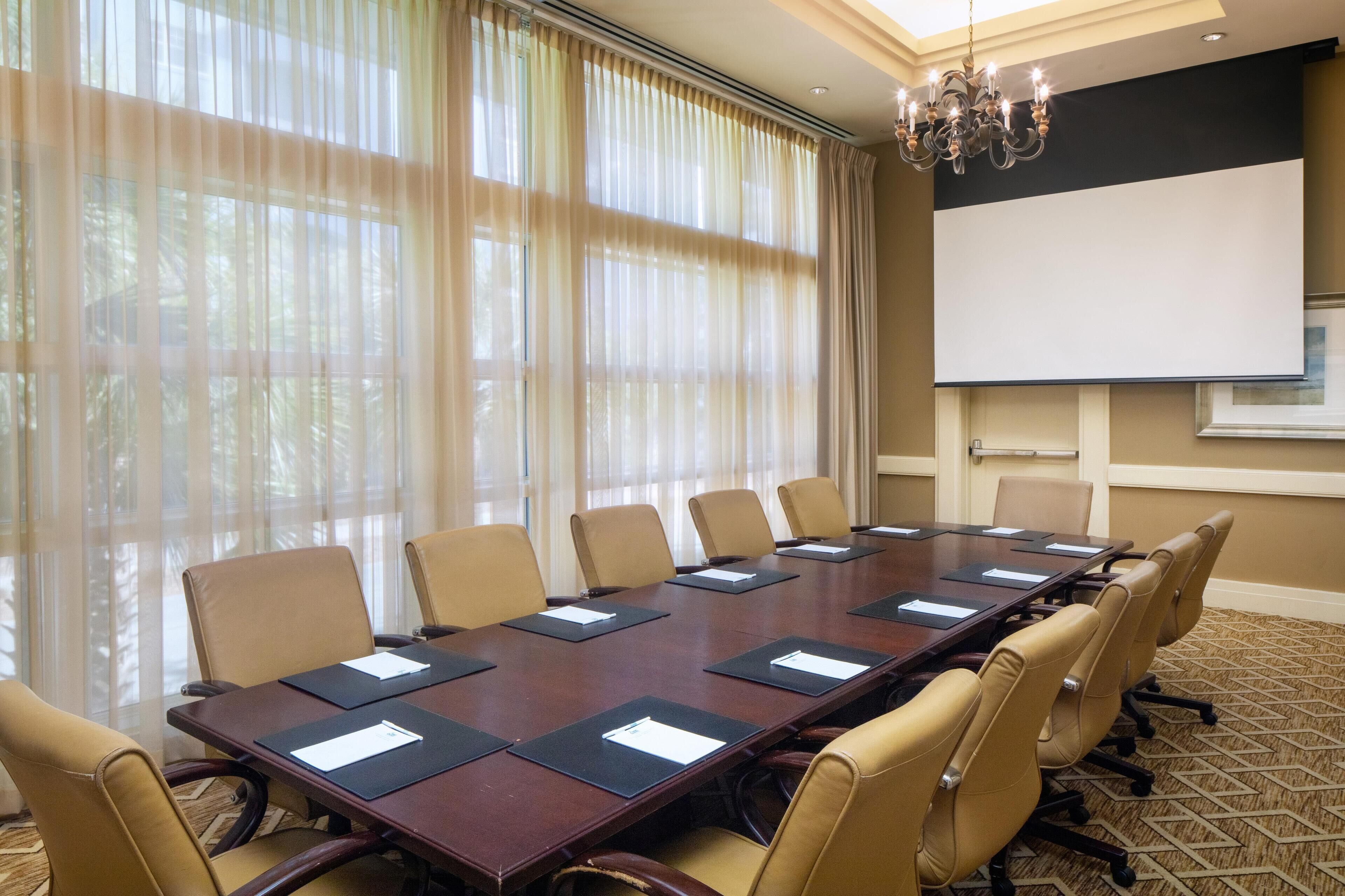 A conference room with a long table, beige chairs, a projector screen, and a chandelier.
