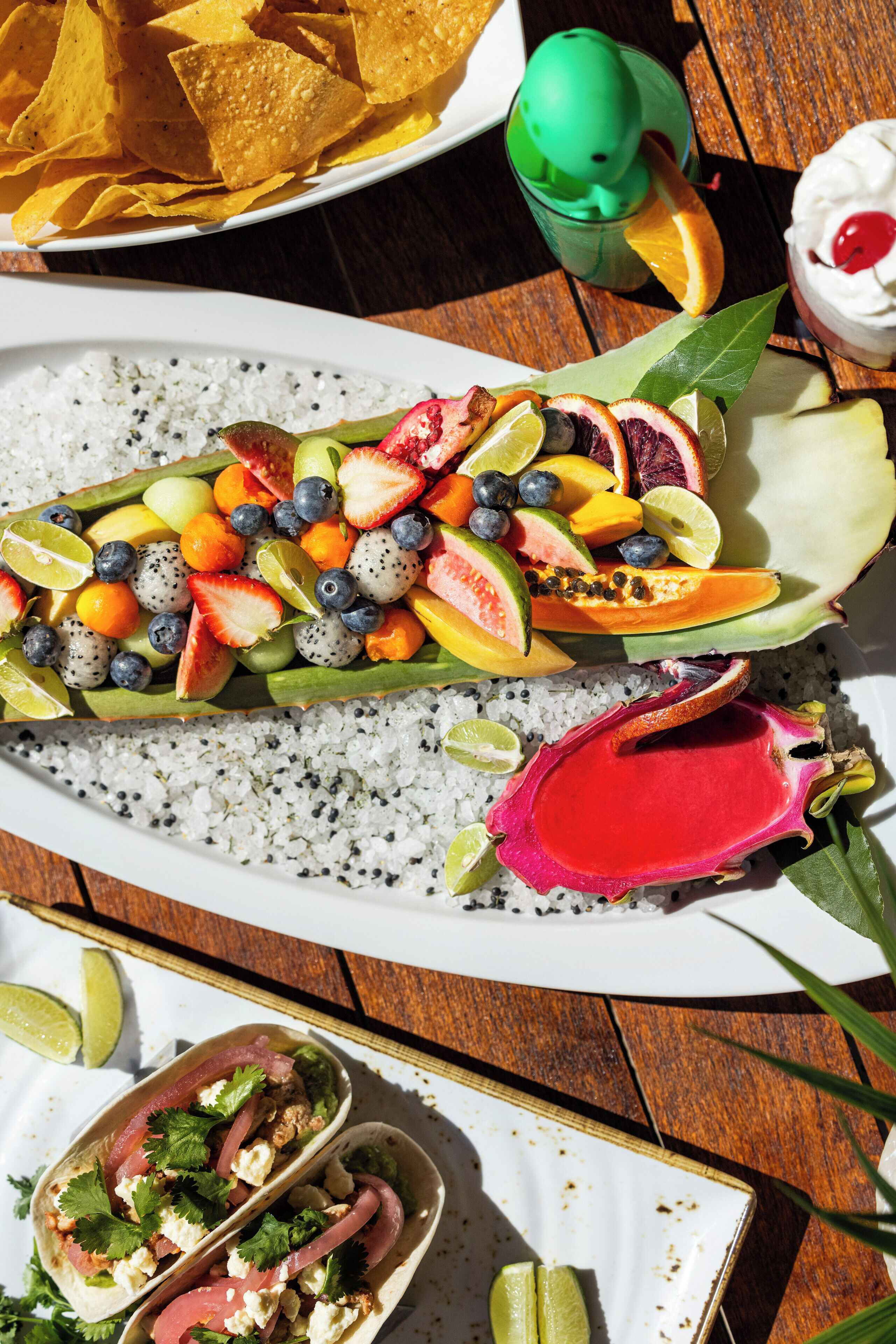 A table with a variety of food items including tacos, chips, and a fruit platter.