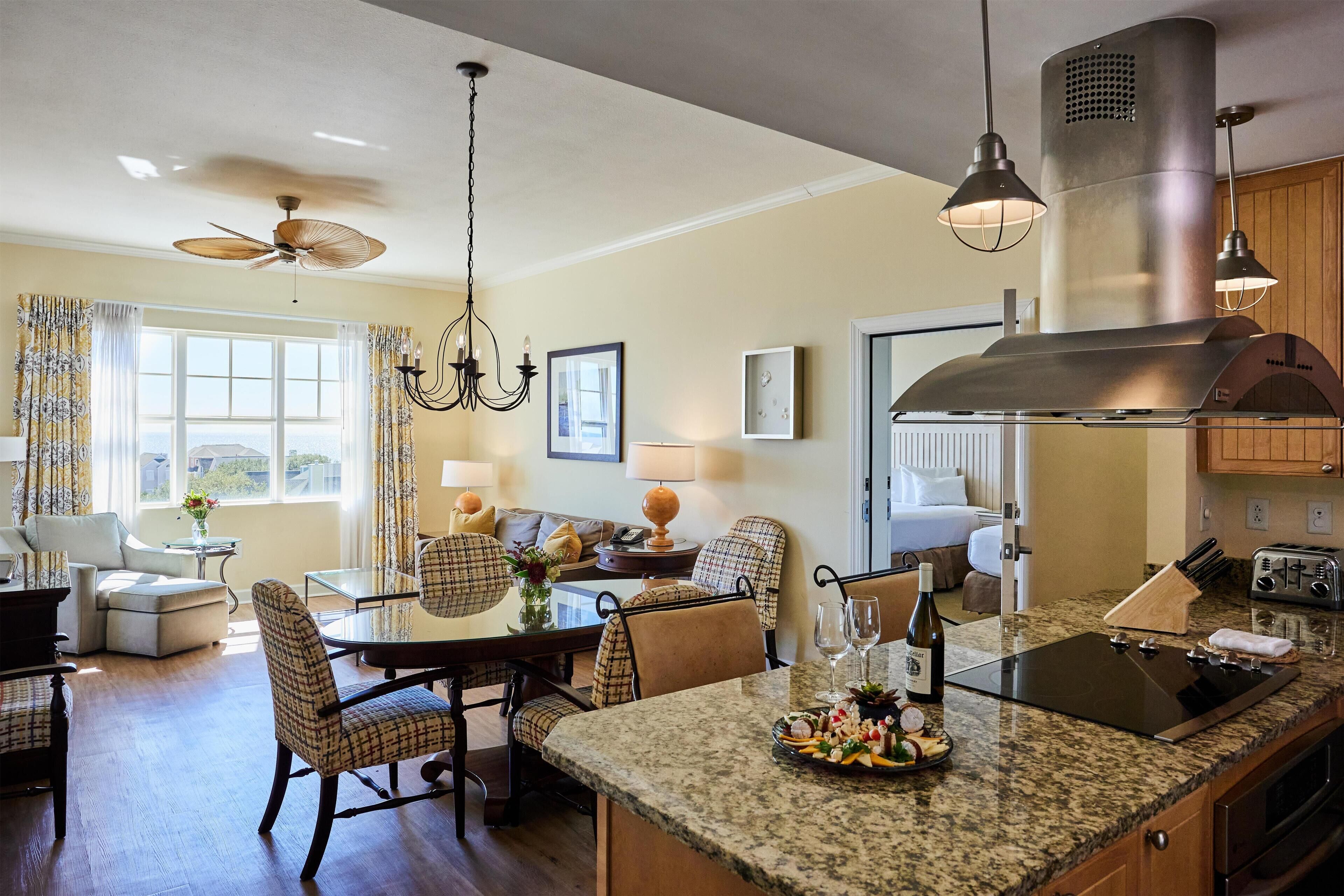 A modern kitchen with granite countertops, a built-in oven, and a stainless steel range hood. There is a dining area with a table and chairs, and a living room with a sofa and a coffee table.