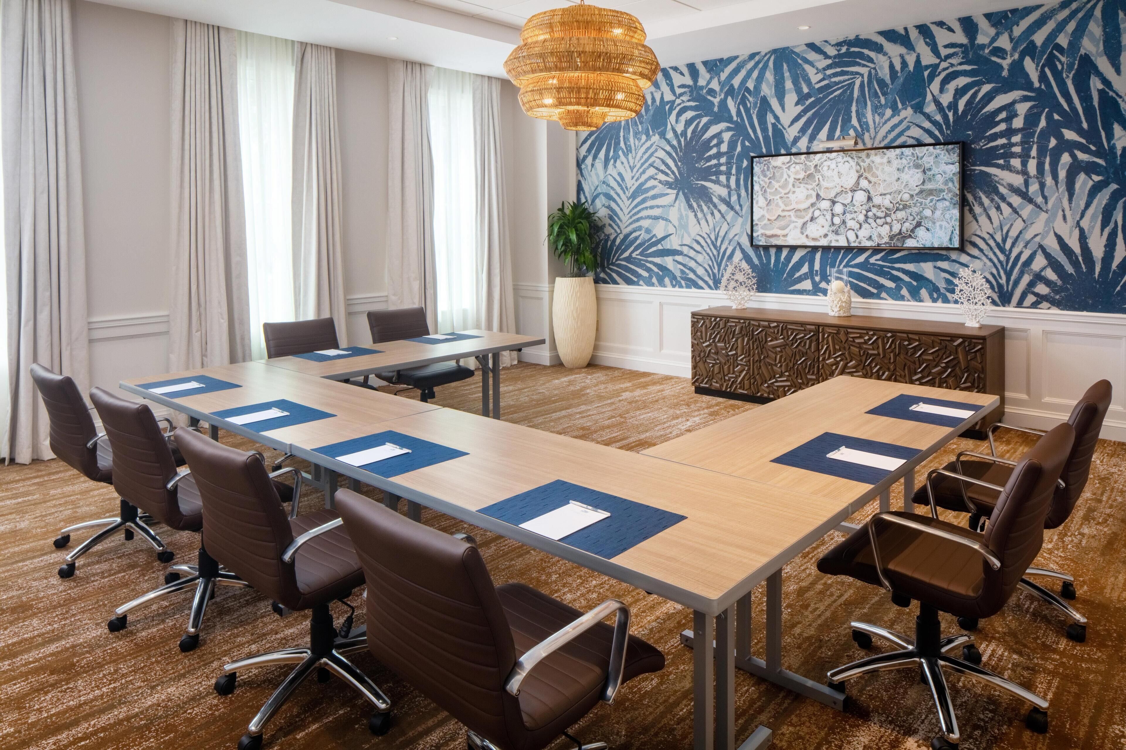 Une salle de conférence avec une longue table, des chaises marron, un mur décoratif et une grande plante.