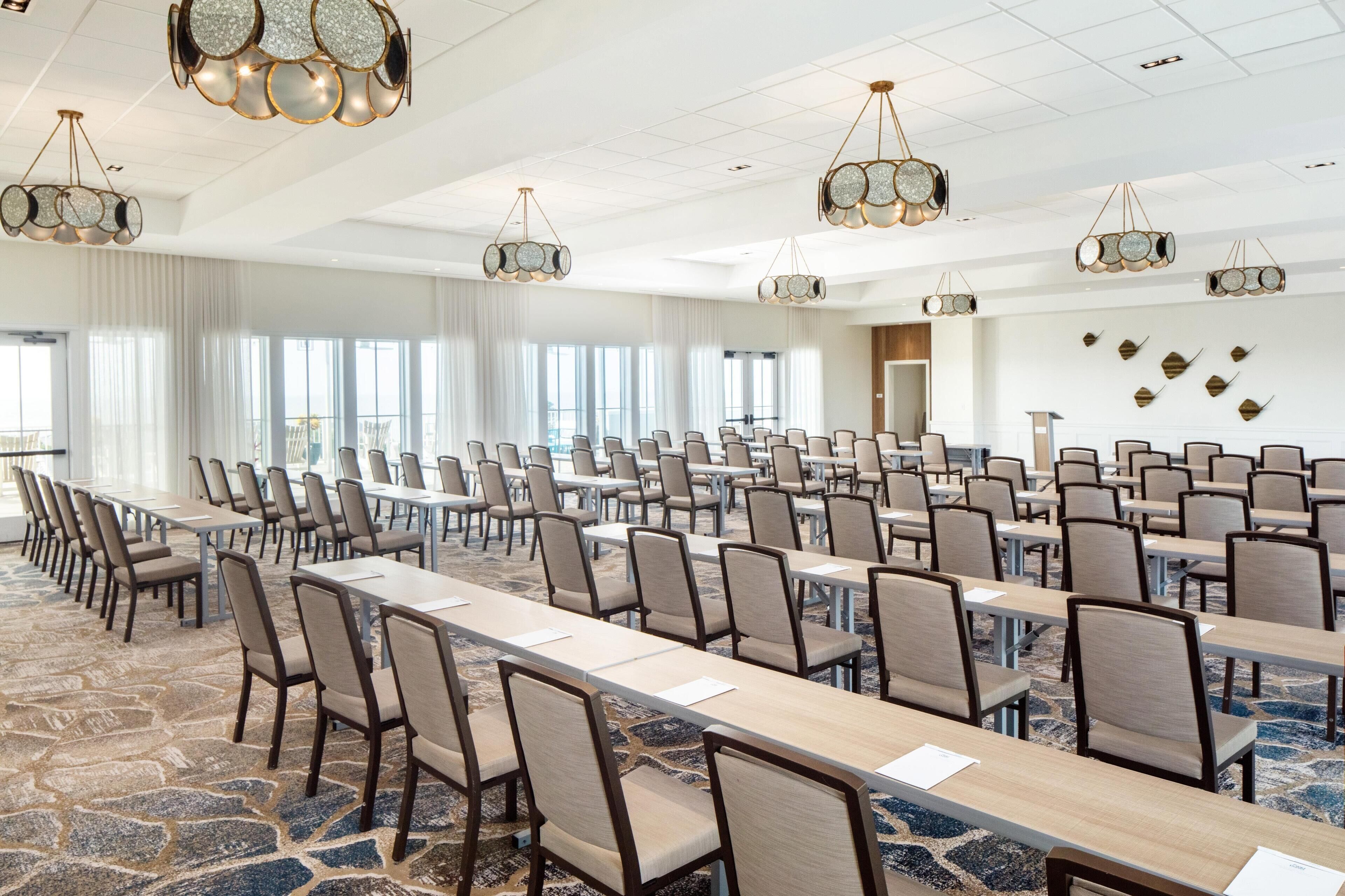 Un salón de conferencias con filas de mesas y sillas, ventanas amplias y elegantes candelabros.