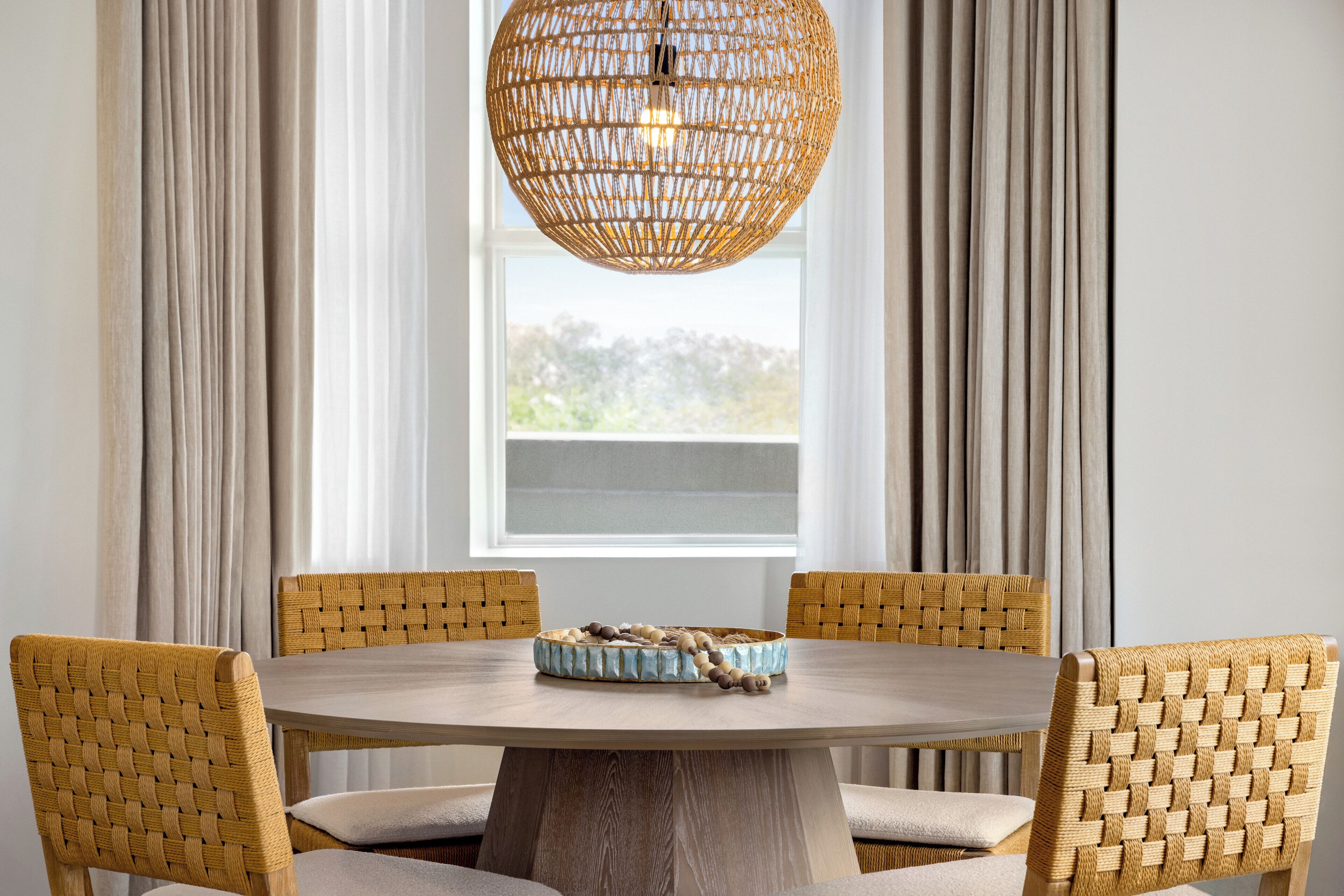A round dining table with a woven chair backrest, a woven pendant light, and a woven bowl on the table.