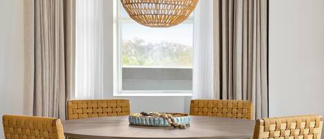 A round dining table with a woven chair backrest, a woven pendant light, and a woven bowl on the table.