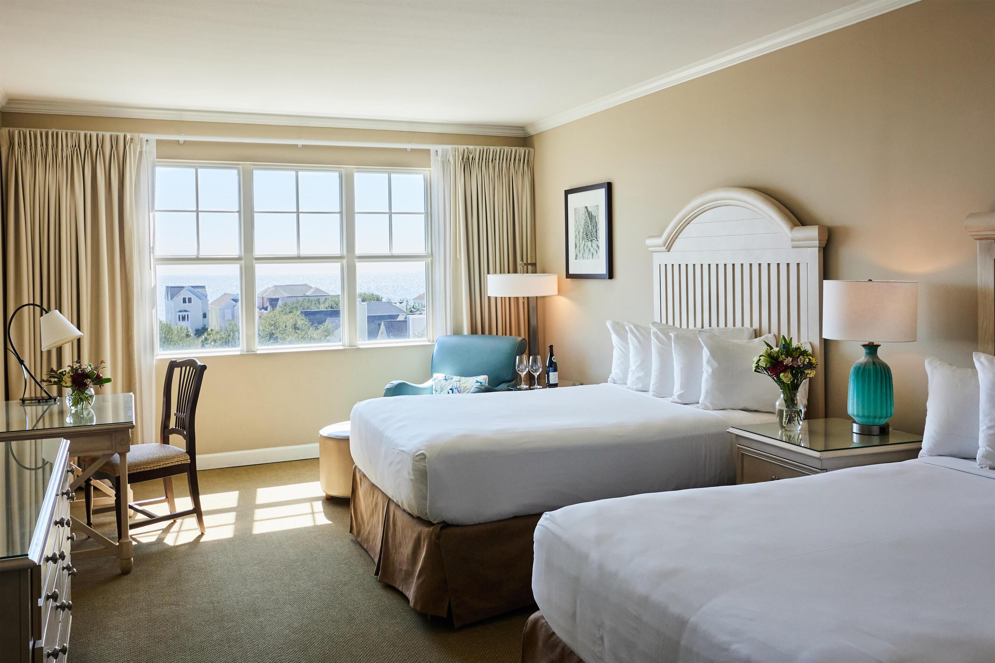 A hotel room with two beds, a desk with a chair, a lamp, and a view of the outside.