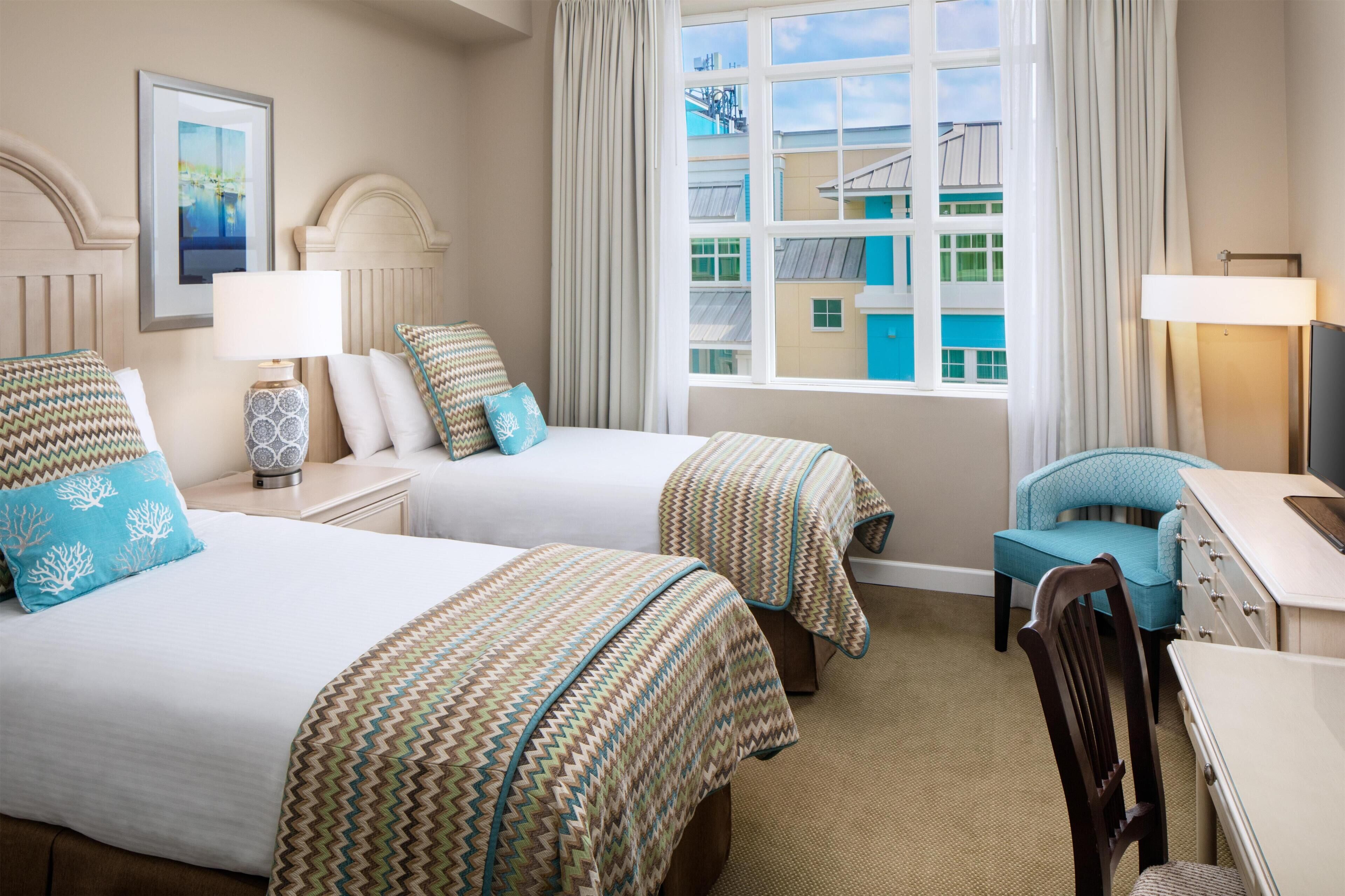 A hotel room with two beds, a desk, a chair, and a window with a view of buildings.