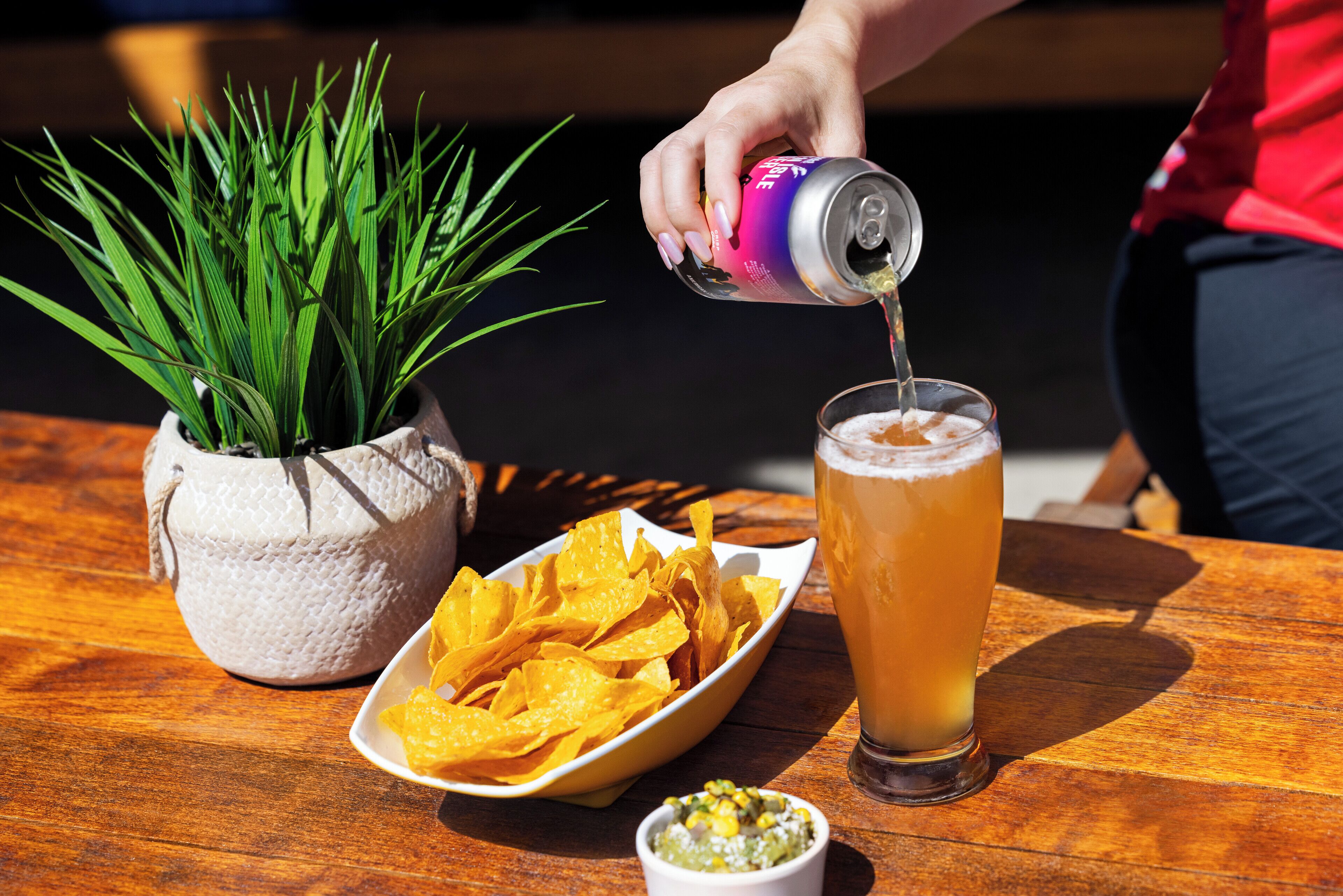 Een hand giet een drankje in een glas, een schaal met chips, een kleine schaal met salsa, en een bloempot op een houten tafel.