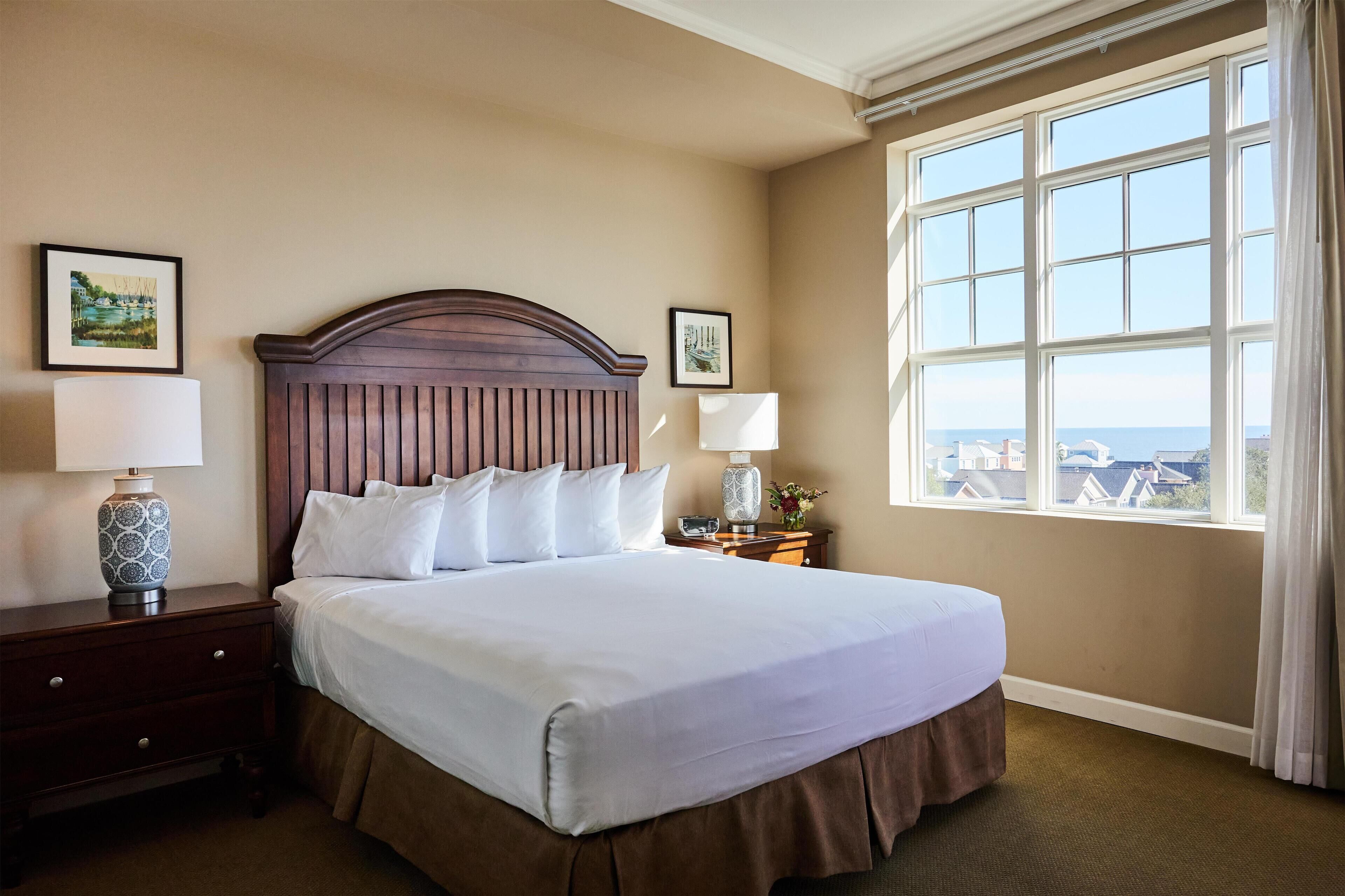 A neatly made bed with a wooden headboard, a nightstand with a lamp, and a window with a view of buildings.