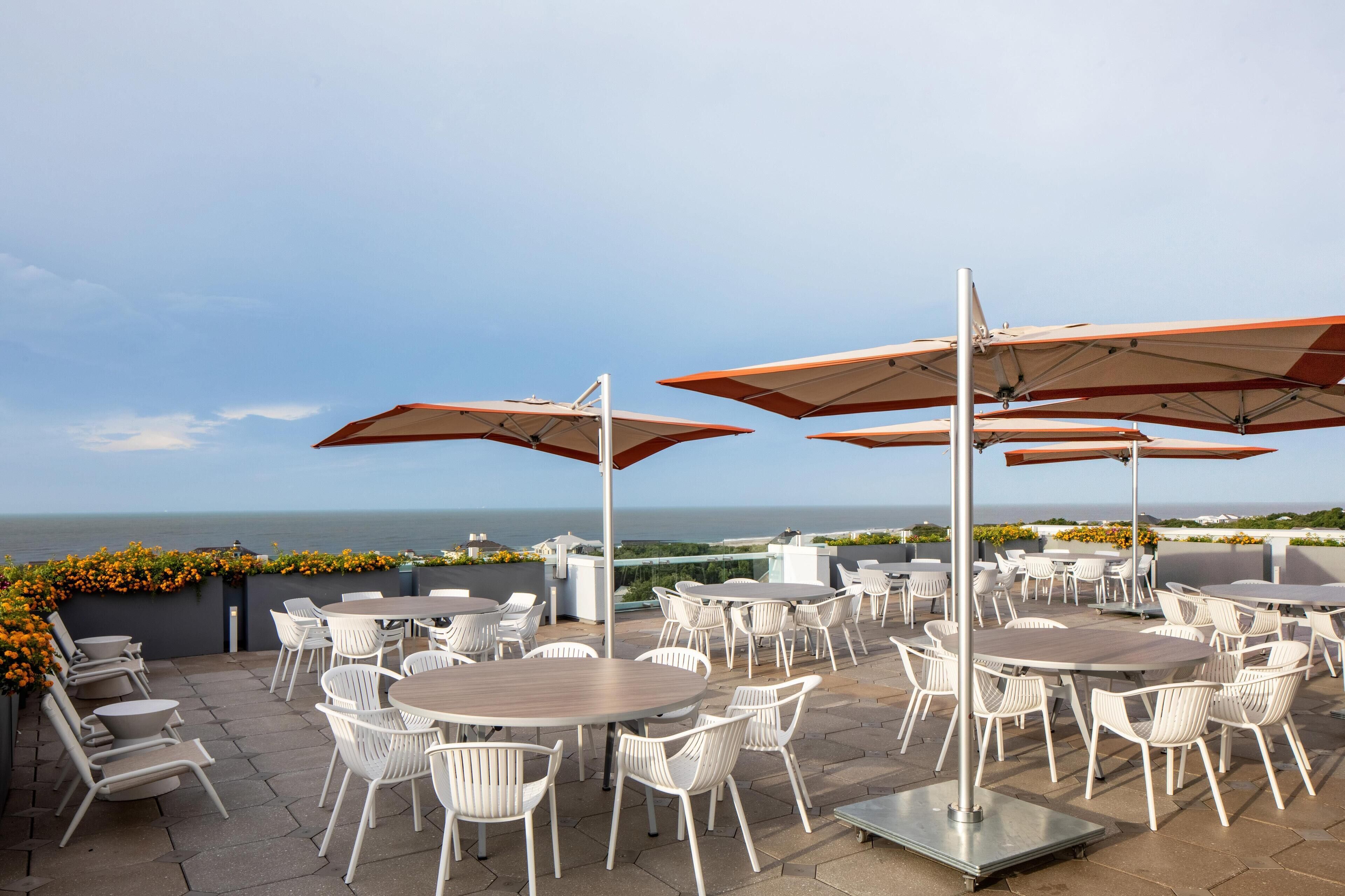 An outdoor patio with white chairs and tables, umbrellas, and a view of the sea.