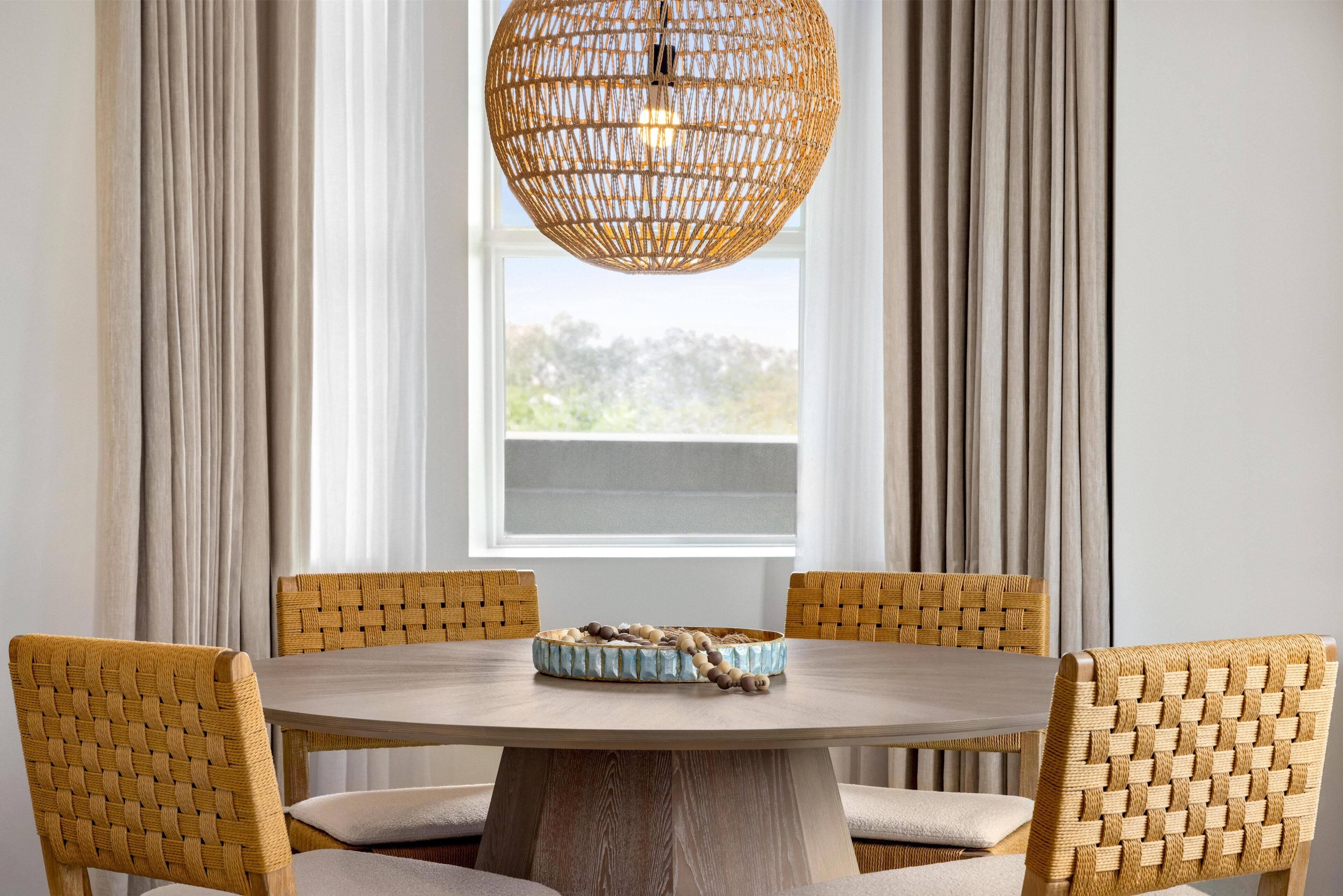 A round dining table with four woven chairs, a woven pendant light, and a woven tray with stones.