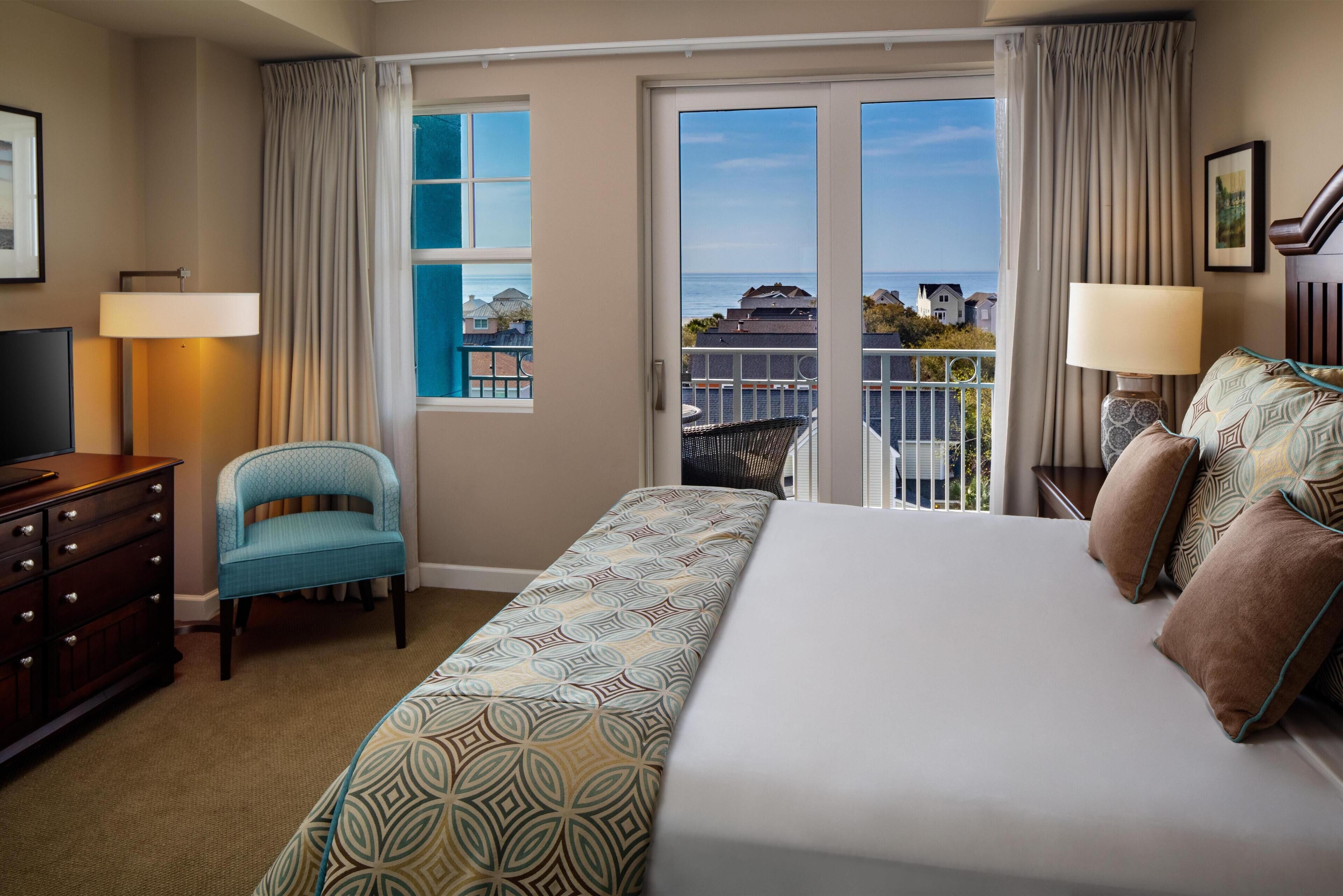 A hotel room with a bed, a chair, a dresser, a TV, and a balcony with a view of the sea.
