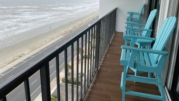 Un balcón con vista a la playa y al océano. Hay sillas turquesas a lo largo de la barandilla.