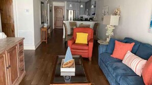Una sala de estar con un sofá azul, un sillón rojo y una mesa de centro con un libro y un gorro de fiesta. Se ve una cocina al fondo.