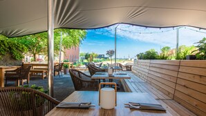 Un espace de restauration en plein air avec des tables et des chaises en bois, un objet cylindrique blanc posé sur l’une des tables, et une vue sur un paysage urbain en arrière-plan.