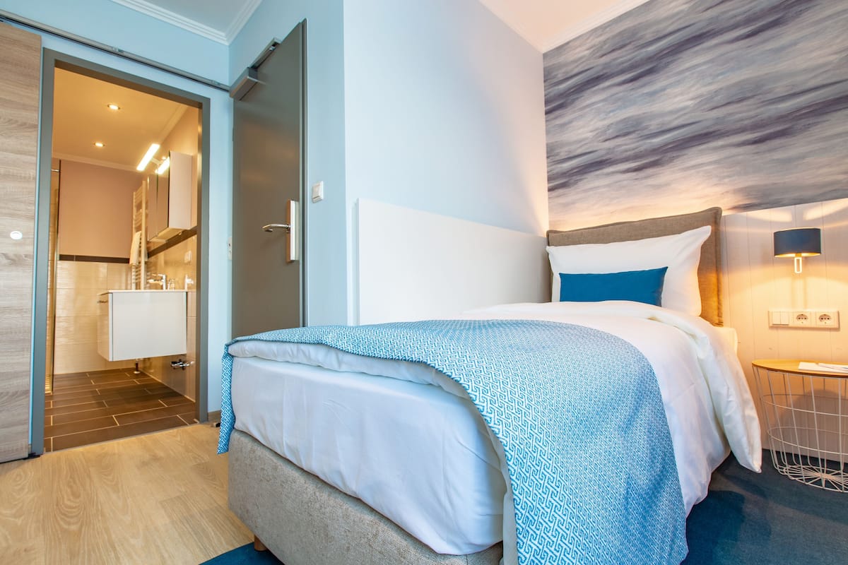 Una habitación de hotel moderna con una cama, una mesita de noche, una lámpara y vistas al baño.