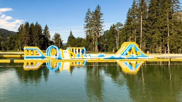 Un parc aquatique doté de structures de jeux gonflables et d’un lac paisible, entouré d’arbres et de collines.