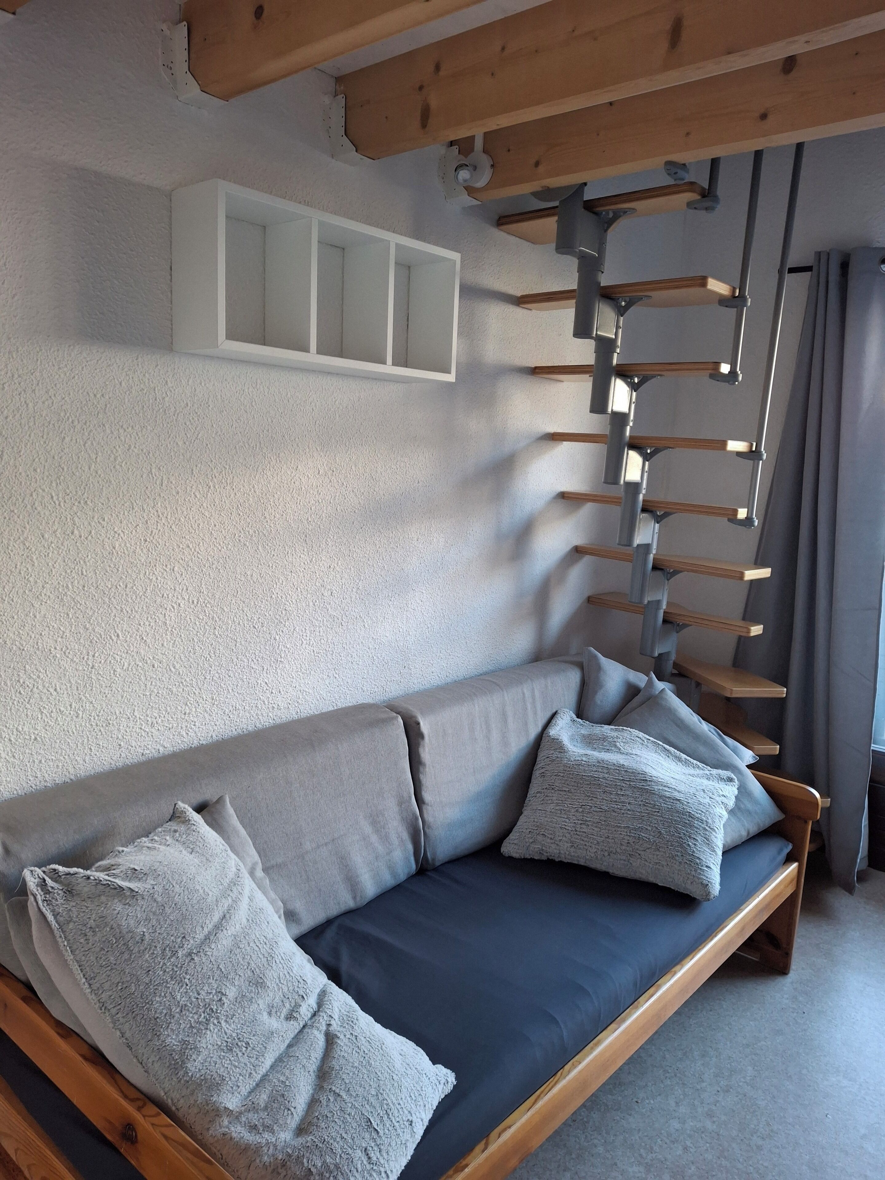 Un salon moderne avec un canapé gris, des poutres en bois et un mur blanc agrémenté d’étagères intégrées.