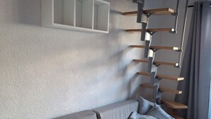 Un salon moderne avec un canapé gris, des poutres en bois et un mur blanc agrémenté d’étagères intégrées.