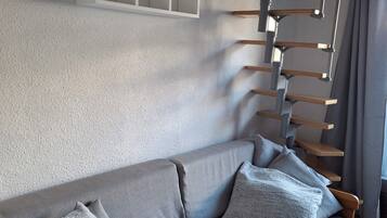 Un salon moderne avec un canapé gris, des poutres en bois et un mur blanc agrémenté d’étagères intégrées.