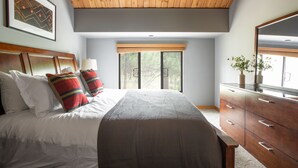 A bedroom with a wooden ceiling, a large bed with white and striped pillows, a dresser, and a window with blinds. - Skylight by AvantStay | Elevated Mountain Home w/ Hot Tub, Billiards,Large Patio (Sunriver)