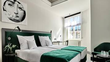 A modern hotel room with a large bed, two bedside tables, a green upholstered chair, and a window with a view of the city.