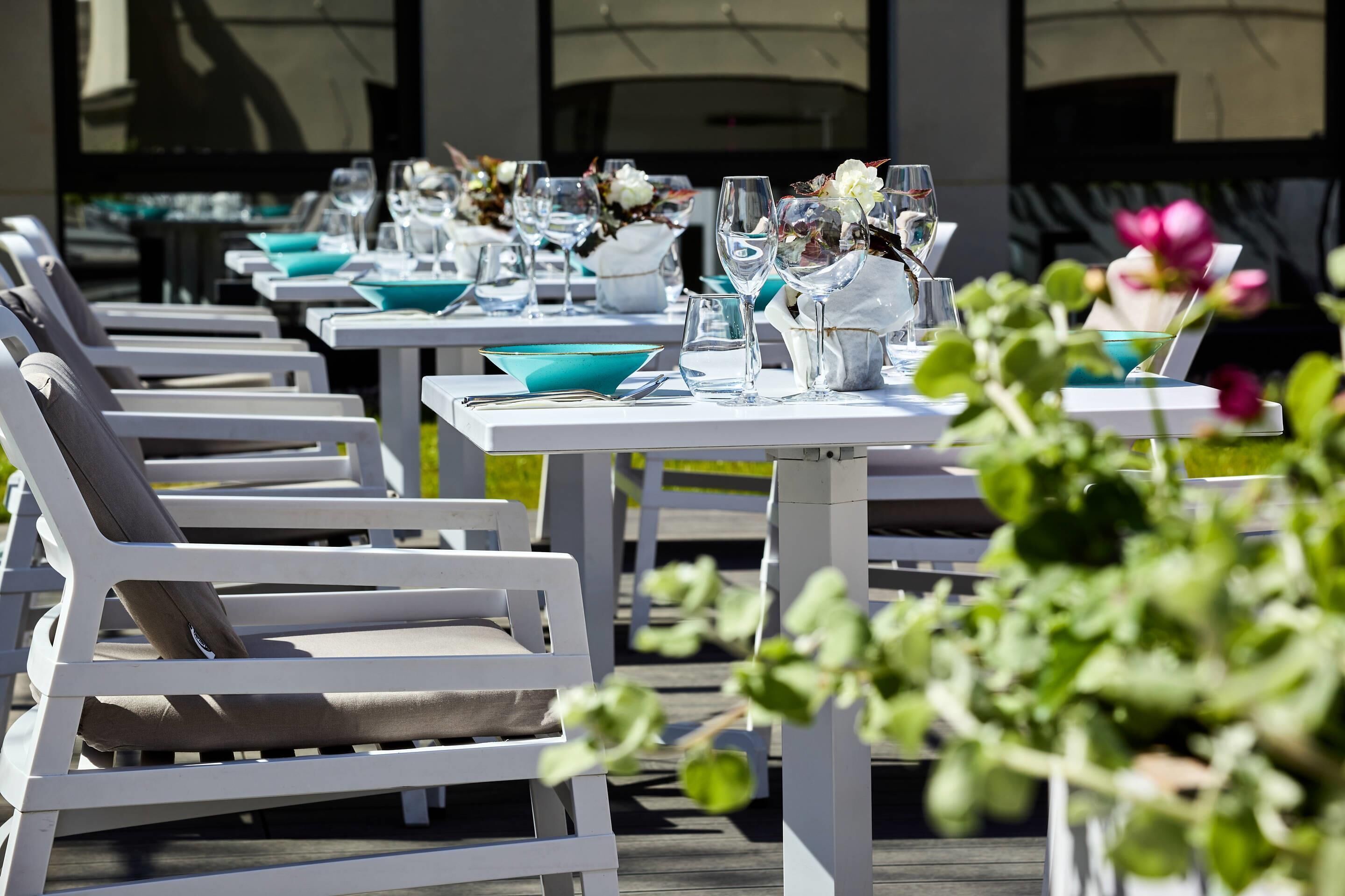 Outdoor dining tables with chairs, each set with a bowl, glassware, and a vase with flowers.