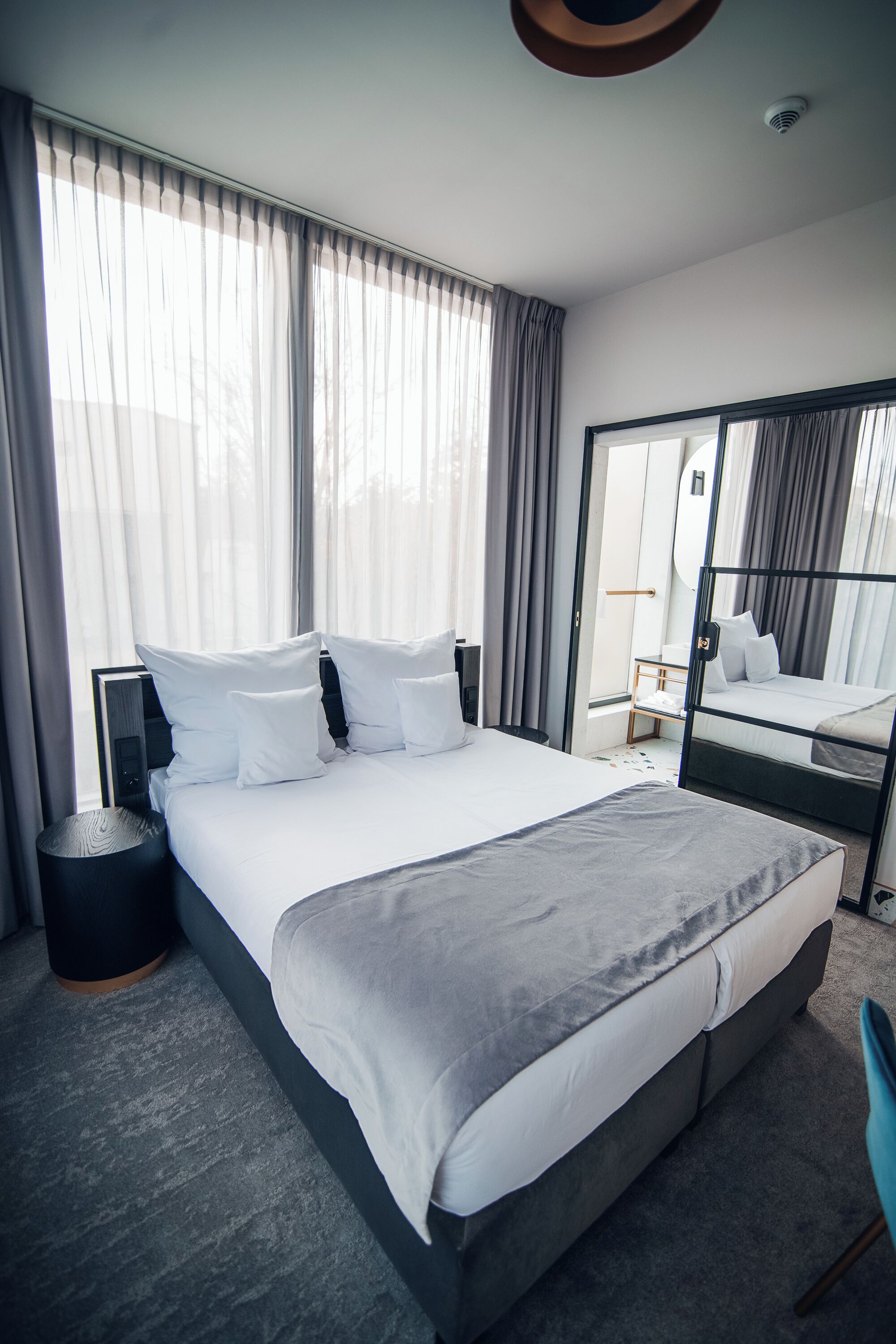 A modern hotel room with a large bed, a bedside table, a chair, a wardrobe, and a mirror.